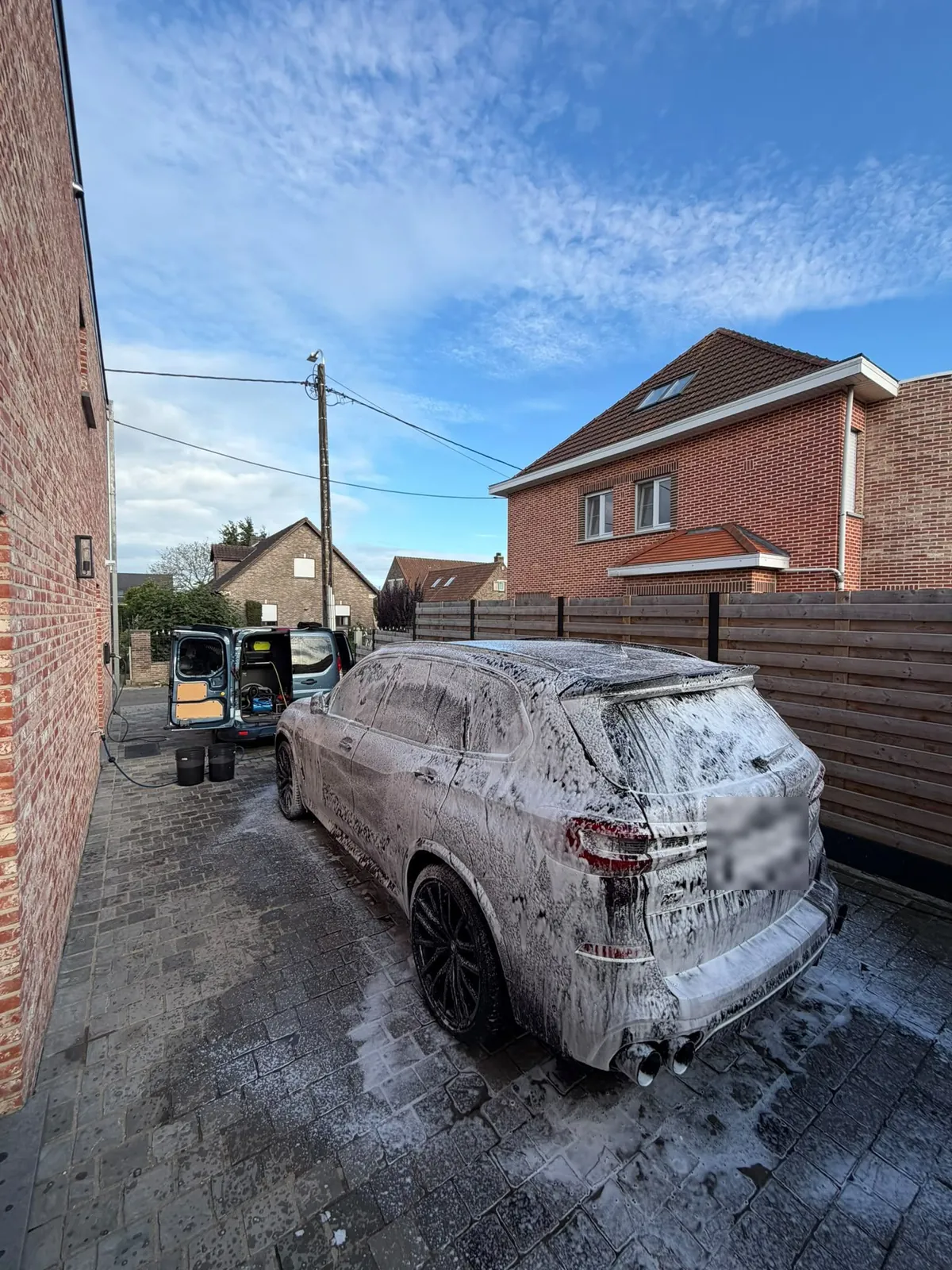BMW X5 onder het schuim op een oprit, met de bus van TB Car Cleaning open op de achtergrond